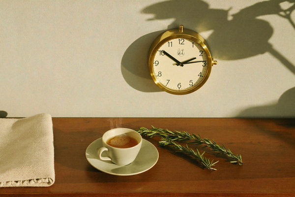 A brass wall clock on a cream wall with tea and rosemary on a walnut shelf