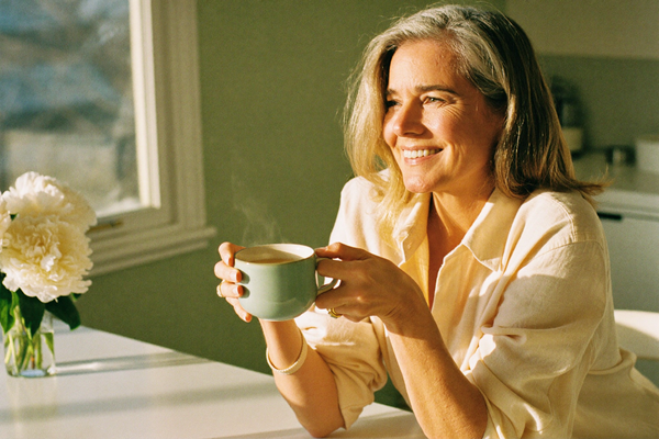 A calm woman holding a warm mug of tea in both hands at a bright kitchen table