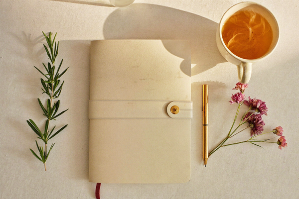 Flat lay of a cream notebook, chamomile tea, and rosemary sprig in warm morning light