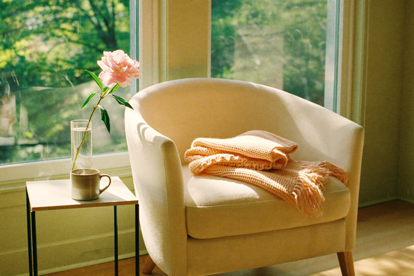 A cream armchair with folded throw beside a sunlit window and peony vase
