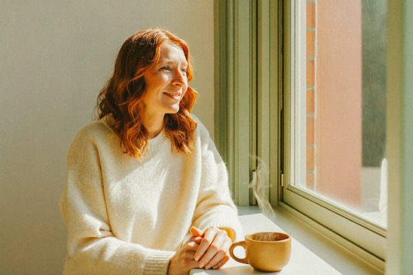 A woman by a bright window with a warm mug of tea, morning light